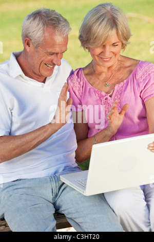 Happy senior man & woman couple sitting together outside in sunshine waving while using a computer for VOIP internet phone call Stock Photo