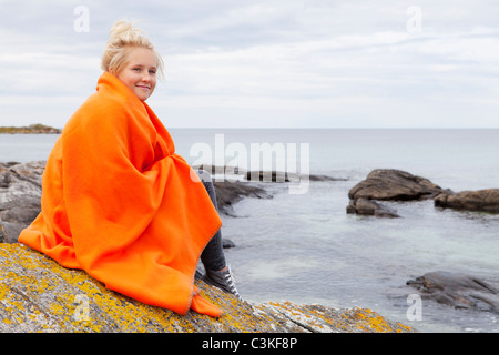 Teenage girl wrapped in blanket sitting by sea, smiling Stock Photo
