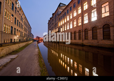 Salts Mill in evening (historic Victorian textile mill building, lights on, reflection on canal water) - Leeds Liverpool Canal, Saltaire, England, UK.