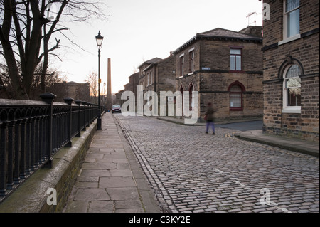 Albert Terrace in historic Saltaire village (end terraced properties, Victorian houses, stone setts on road, mill chimney) - Bradford, Yorkshire, UK. Stock Photo