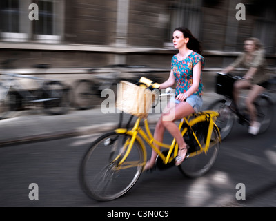 Cyclists in Cambridge. Two girls in shorts riding through streets against backdrop of university buildings. Striking yellow bike Stock Photo