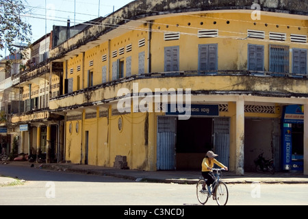 A young Asian girl is riding a bicycle past a colonial style building on a street near the Mekong River in rural Kratie, Cambodi Stock Photo
