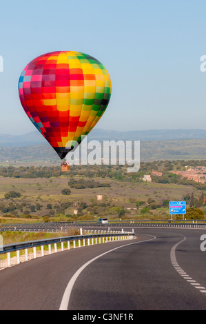 A multicoloured hot air balloon just about landing dangerously on a motorway.  Hills and a blue sky in the background. Stock Photo