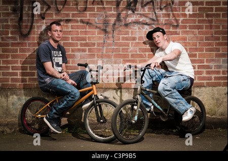 Two teenage boys with attitude on BMX stunt bikes leaning against a brick wall, UK Stock Photo