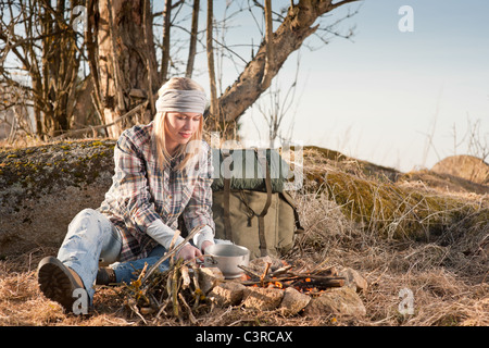 Young hiking woman with backpack cook on campfire in countryside Stock Photo