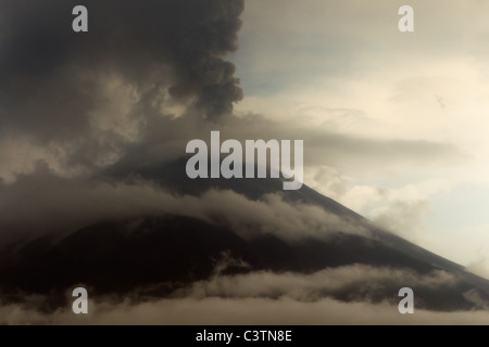 A large volcanic mountain is partially shrouded in clouds, with a thick, dark plume of ash and smoke erupting from its summit and rising into the overcast sky. The mountain's slopes are dark and Stock Photo