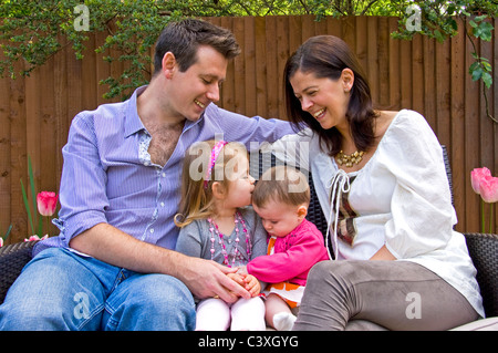 Horizontal family portrait of an attractive husband and wife laughing as one daughter gives the other a kiss on the head. Stock Photo