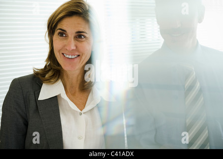 Smiling businesswoman looking away, businessman behind glass wall Stock Photo