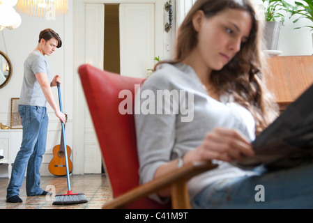 Man sweeping floor while wife reads Stock Photo