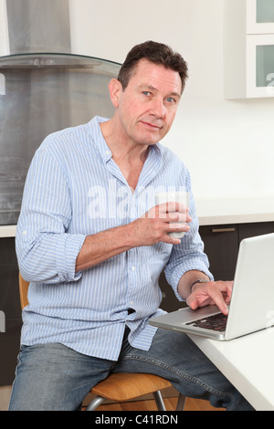 Man with tea or coffee in hand sitting at home in the kitchen using a laptop, looking at camera Stock Photo