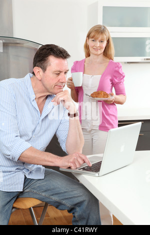 Man sitting using a laptop at home, woman holding a mug of tea.coffee looking on. Stock Photo