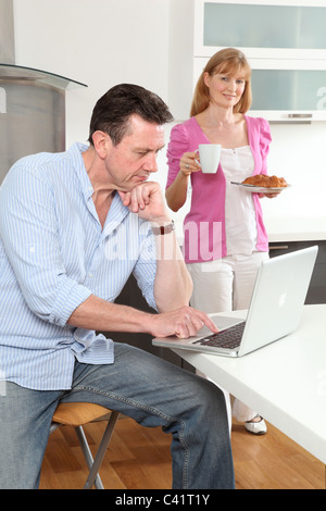 Man sitting using a laptop at home, woman holding a mug of tea.coffee looking on. Stock Photo