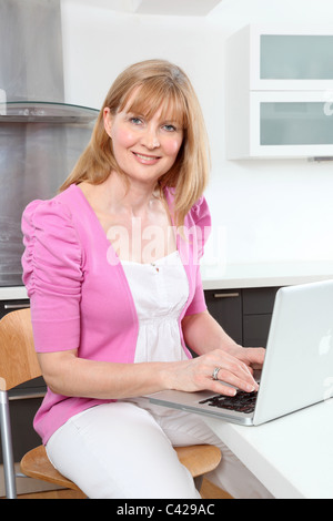 Smiling woman sitting in a kitchen using a laptop. Stock Photo