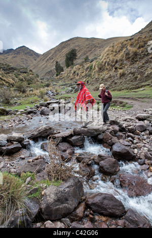 Village near Ollantaytambo. Indian man in traditional dress and tourist, woman, crossing stream druing hike. Stock Photo
