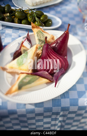 Cheese toast triangles and sliced pickled pears on a paper plate with green olives in the background Stock Photo