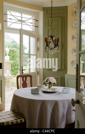 Dining table with tablecloth in country style kitchen with chandelier, decorative plates and view through back door to garden Stock Photo