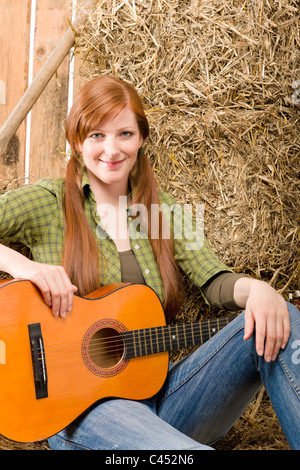 Young country woman sitting on hay with guitar in barn Stock Photo