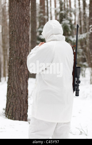 Soldier in white camouflage aiming with sniper rifle at winter forest. Stock Photo