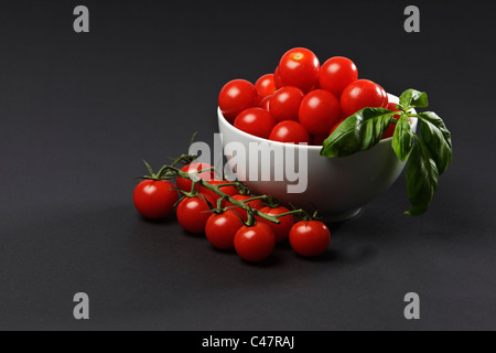 A white bowl of cheery tomatoes with a sprig of basil against a plain black background Stock Photo