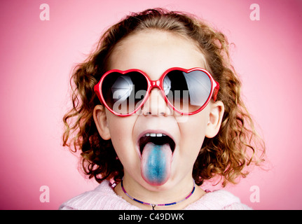 A young girl with curly hair on a pink studio wall wearing heart-shaped sunglasses and sticking out a stained blue tongue Stock Photo