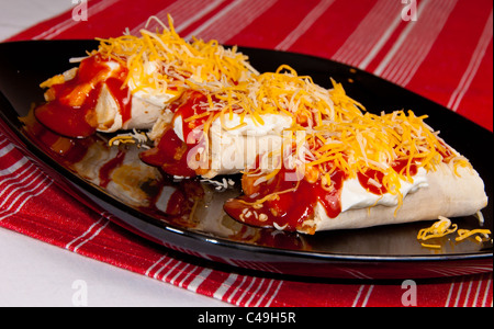 Three Burritos covered in Sour Cream, Enchilada Sauce, and Shredded Cheese on a Black Plate on a Red Cloth. Stock Photo