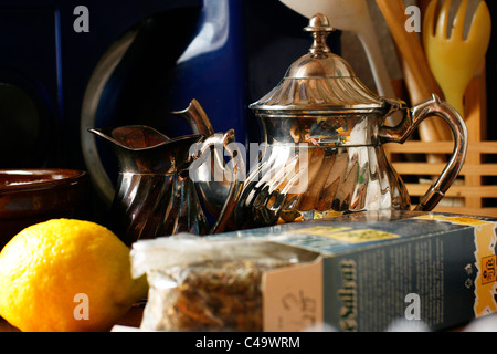 Herbal tea, an old silver teapot, a small milk jug and a lemon in front of other kitchen stuff Stock Photo