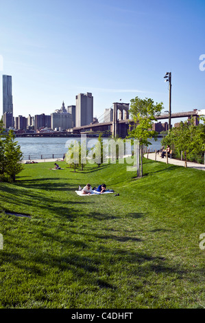 people enjoy relaxing on grassy sloping lawn in Brooklyn Bridge park overlooking esplanade East River & Lower Manhattan skyline Stock Photo