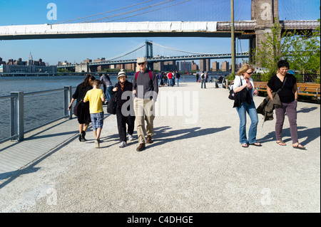 people enjoy strolling on Brooklyn Bridge park esplanade overlooking East River  next to section of bridge under repair Stock Photo