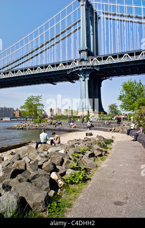 people sit reading relaxing sunning on stone bleachers at waters edge in area of Brooklyn Bridge park next to Manhattan bridge Stock Photo