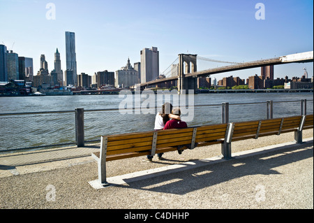 solitary courting couple sit on bench of Brooklyn Bridge park esplanade  overlooking East river & lower Manhattan skyline Stock Photo