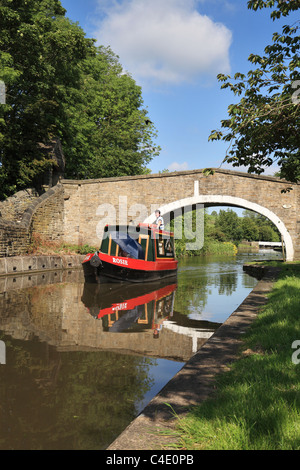 A canal day hire narrowboat passes under a stone bridge Kildwick, North Yorkshire, England, UK Stock Photo