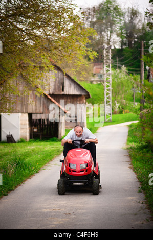 Cheerful gardener riding tractor mower on countryside road. Shallow DOF Stock Photo