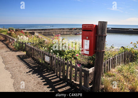 A red post box with Craster Harbour in the background. Northumberland, North EastEngland, UK Stock Photo
