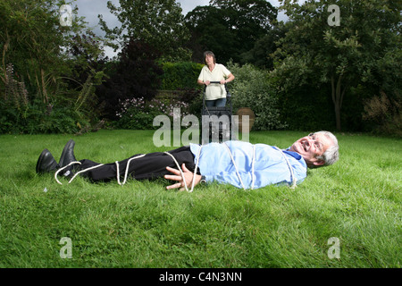 senior woman pushing lawn mower towards tied up husband Stock Photo