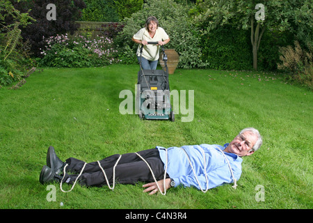 senior woman pushing lawn mower towards tied up husband Stock Photo