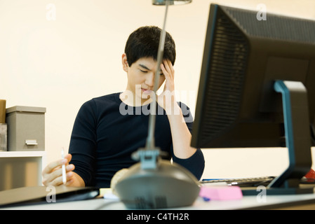 Young man sitting at desk, holding head and looking down in thought Stock Photo
