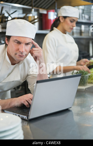 USA, New York, New York City, Chef using laptop and cook preparing food in commercial kitchen Stock Photo
