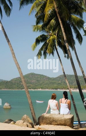 Two young women sit among palm trees high over the beach at Palolem Beach, in Goa state, India Stock Photo