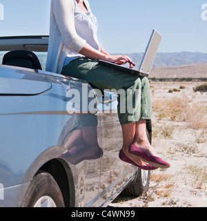 USA, California, Palm Springs, Woman sitting on side of convertible car in desert, using laptop Stock Photo