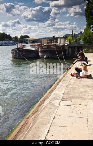 young woman female student in Paris France relaxing beside river Seine with book in sunshine river barges dramatic blue sky cumulus clouds background Stock Photo