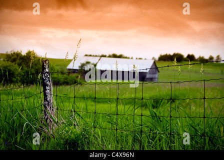 An old, rotting wire fence stands in the foreground with an old barn in the background, under a stormy, orange sky. Stock Photo
