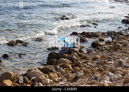 Man sat under a umbrella on the rocks looking out at the ocean, View of Pafos Cyprus Stock Photo