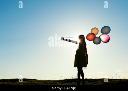 Silhouette of a young girl holding coloured balloons at sunset