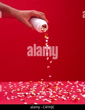 A man's hand holding a container while poring pills and tablets on a scattered red surface Stock Photo