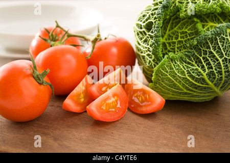whole and cut tomatoes with cabbage on wooden board. White bowl and plate in the background. Stock Photo
