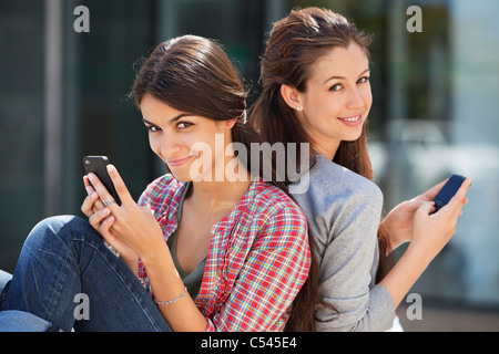 Portrait of two young women sitting back to back and using mobile phones Stock Photo
