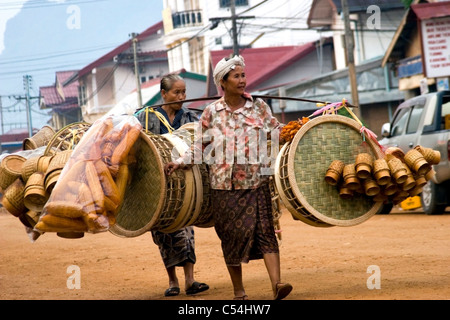 Two Asian women are carrying large loads of assorted baskets on a dirt street in communist Laos. Stock Photo
