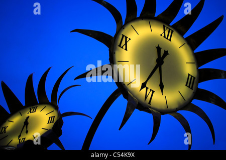 The bright yellow faces of two clocks resembling sunflowers and shoot at twilight are in sharp contrast against a deep blue sky. Stock Photo