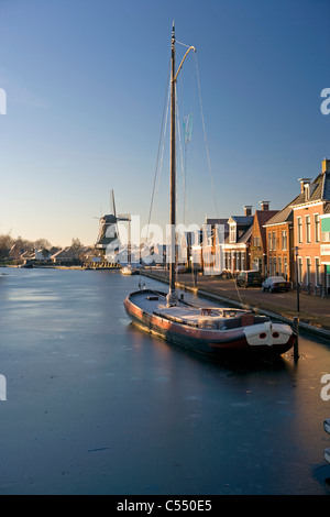 The Netherlands, Woudsend, Traditional cargo ship, now sailing boat for tourism in frozen canal. Background windmill. Stock Photo