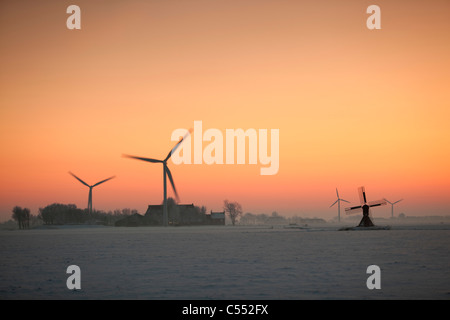 The Netherlands, Tritzum, Small traditional windmill, wind turbines and farm in snow at sunset. Stock Photo